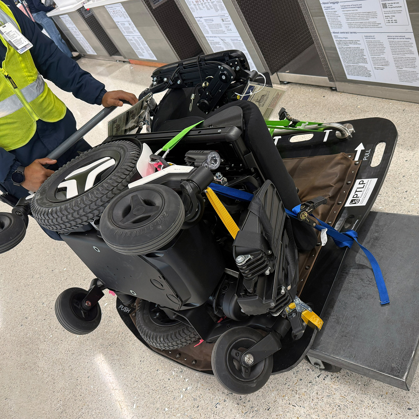 A powered wheelchair is laying on its side and strapped to the Padded Tipping Plate, which rests on a small airport cart pushed by an air carrier agent.
