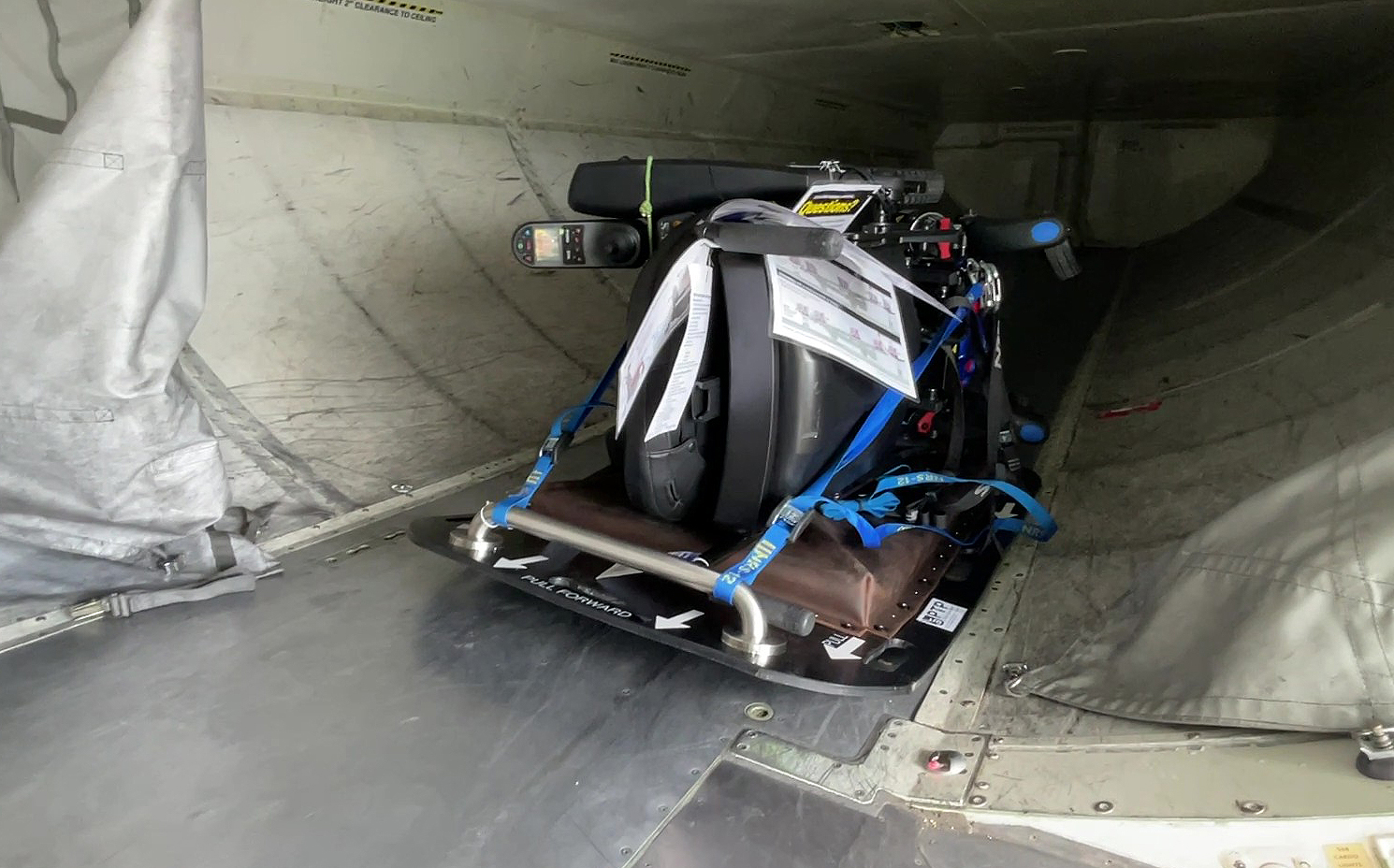 Tipped powered wheelchair and padded tipping plate package lying on the floor of an aircraft cargo hold.